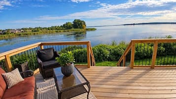 A deck with a sofa, a table, and a potted plant, overlooking a calm lake with a distant shoreline.