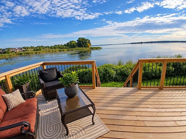 A deck with a sofa, a table, and a potted plant, overlooking a calm lake with a distant shoreline. - Pet friendly Waterfront Stay Near Wineries, Beaches & Trails with kayaks (Quinte West)