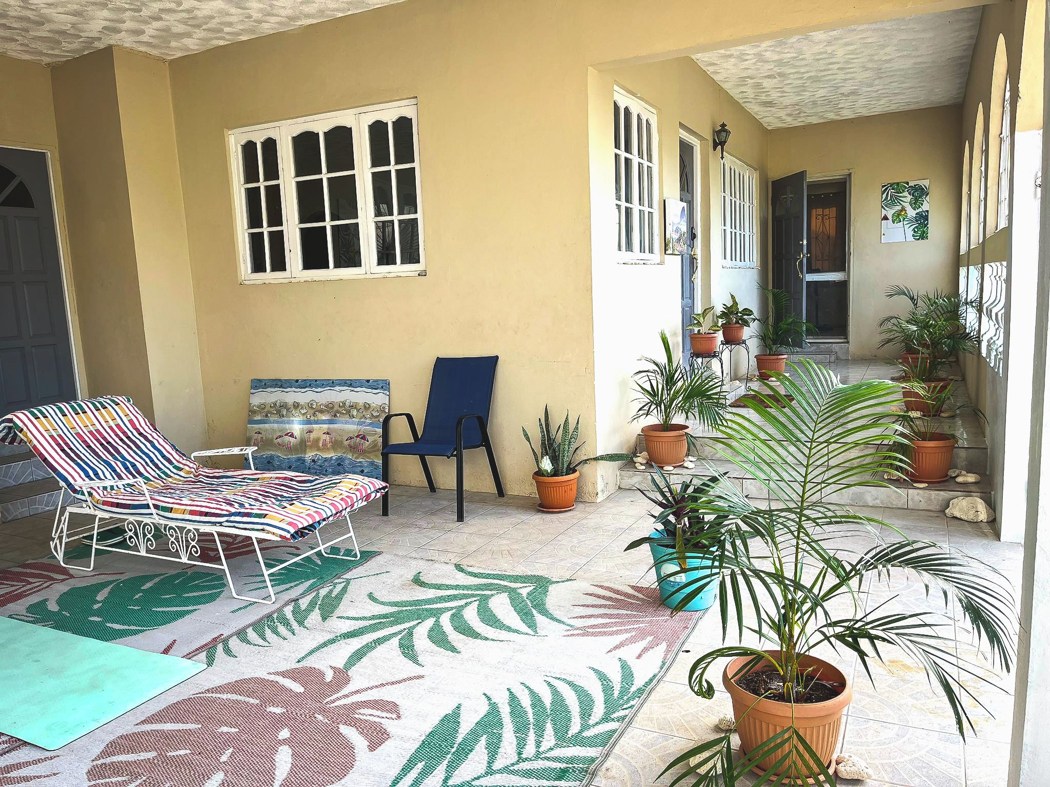 A covered outdoor area with a lounge chair, a blue chair, and potted plants.