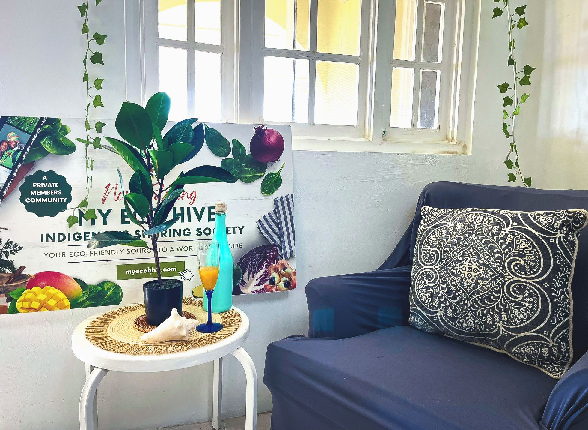 A room with a dark blue sofa, a small white table with a plant, a bottle, and a glass of orange juice. There's a poster on the wall with text and images of fruits and vegetables.