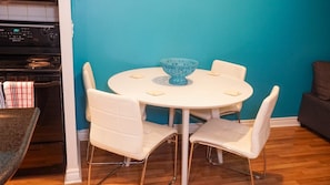 A dining area with a round table and four chairs, a blue bowl on the table, and a kitchen with a stove in the background. - Margaritaville (Toronto)