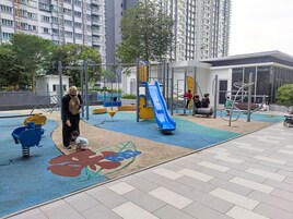 A playground with a slide and climbing structures, surrounded by a paved area and tall residential buildings.