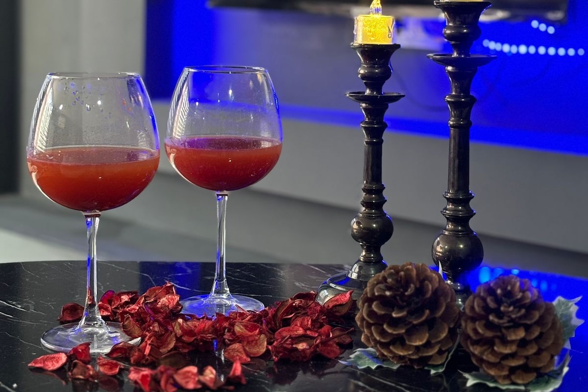 Two glasses of red liquid on a reflective surface with decorative pine cones and dried flowers, a candle holder with a candle, and a television in the background.