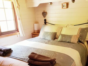 A neatly made bed with pillows and a blanket, a bedside table with a lamp, and a window with curtains. - River Lodge (Lynton)