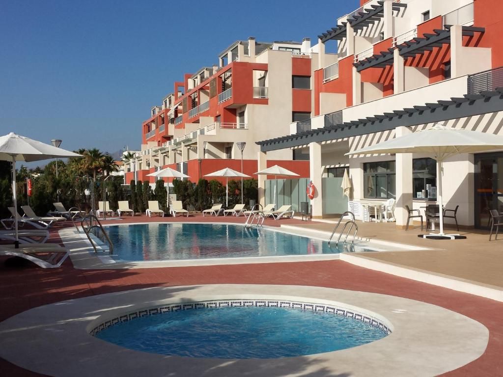 Zona de piscina con sillones de playa y sombrillas, rodeada por un edificio de varios pisos con balcones.
