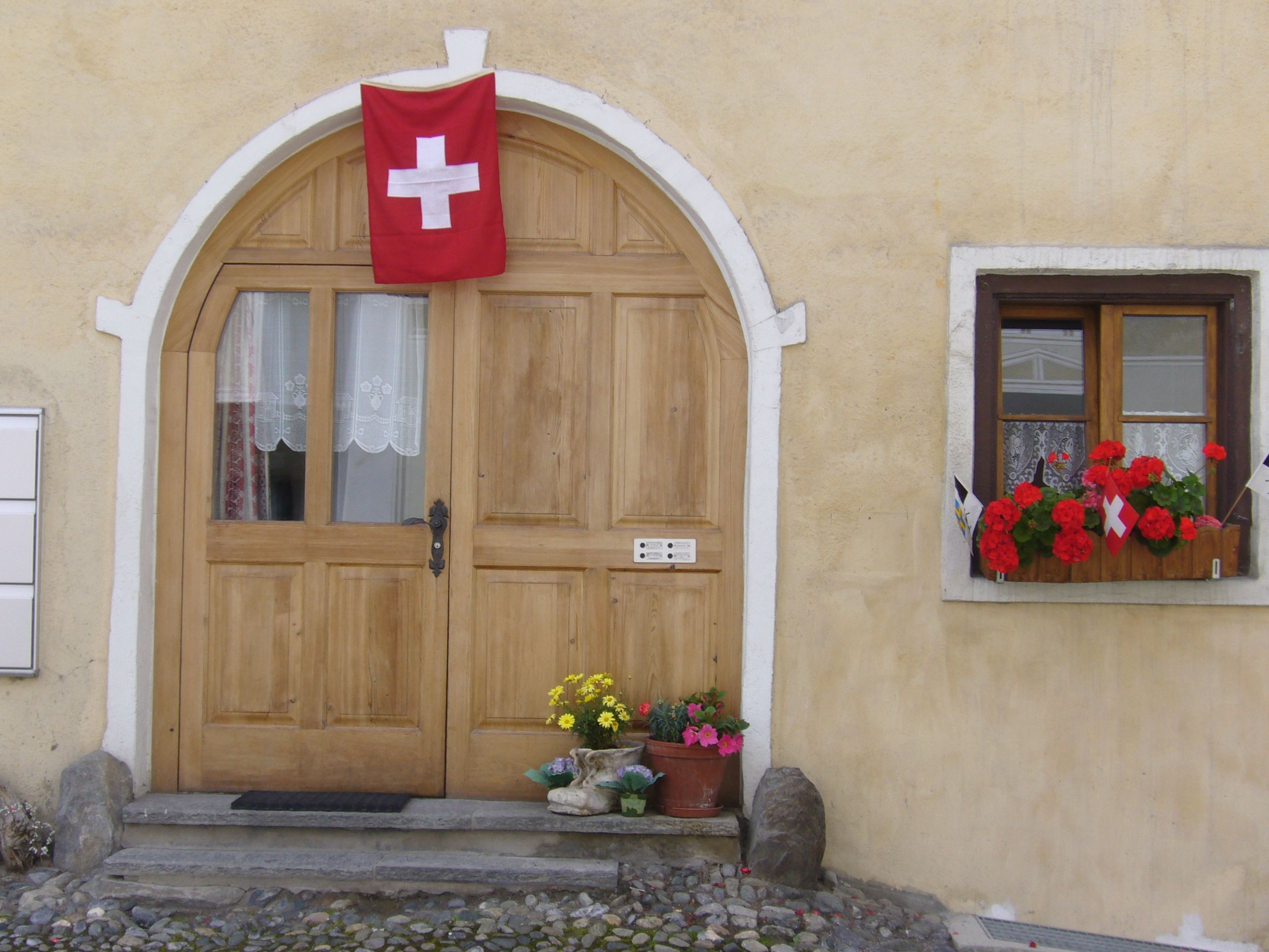 Eine Holztür mit einer roten Fahne, ein Fenster mit Blumen und ein Steinweg, der zur Tür führt.