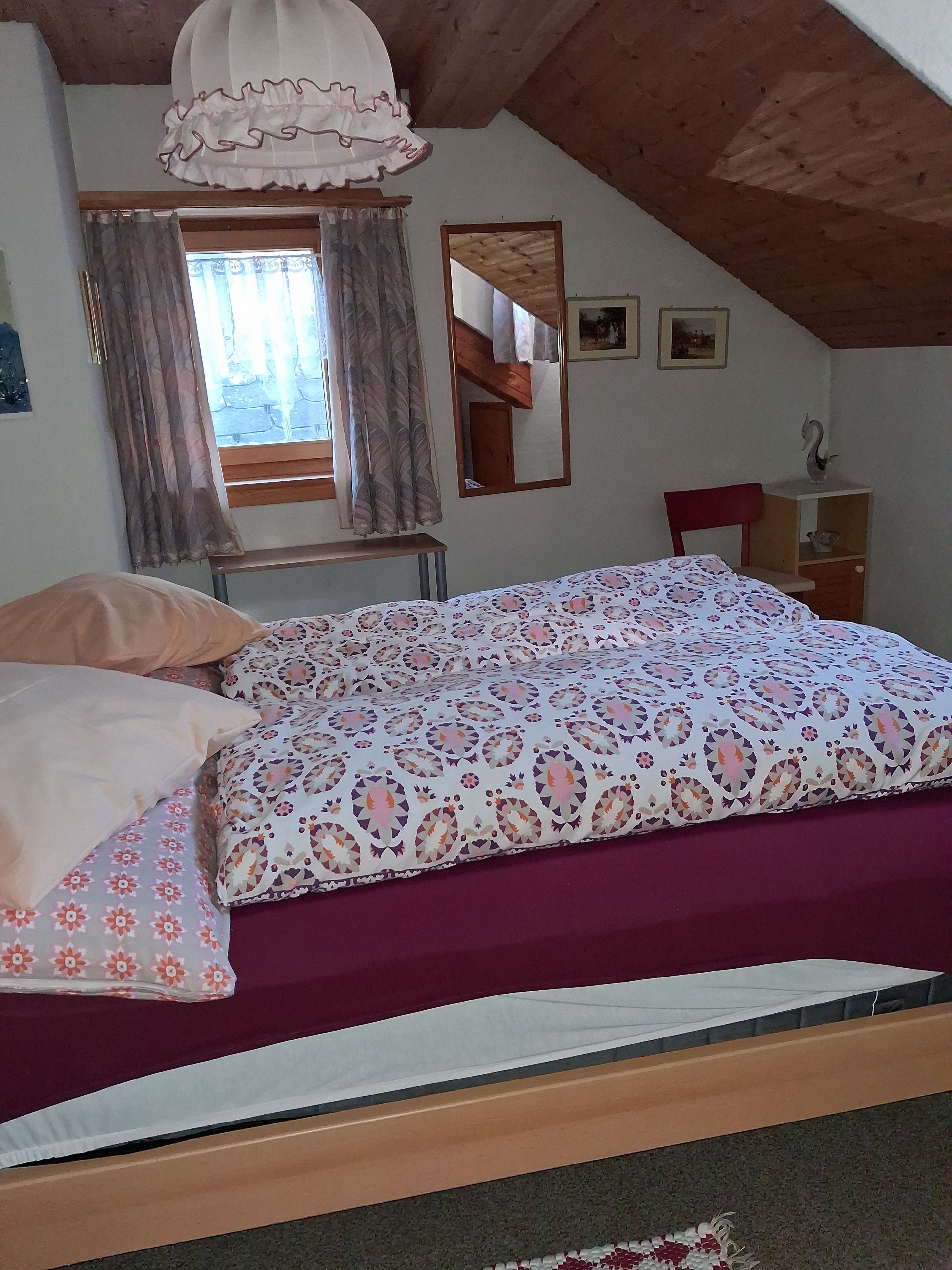 A bedroom with a bed, a window with curtains, a mirror, and a small wooden shelf with a swan figurine.