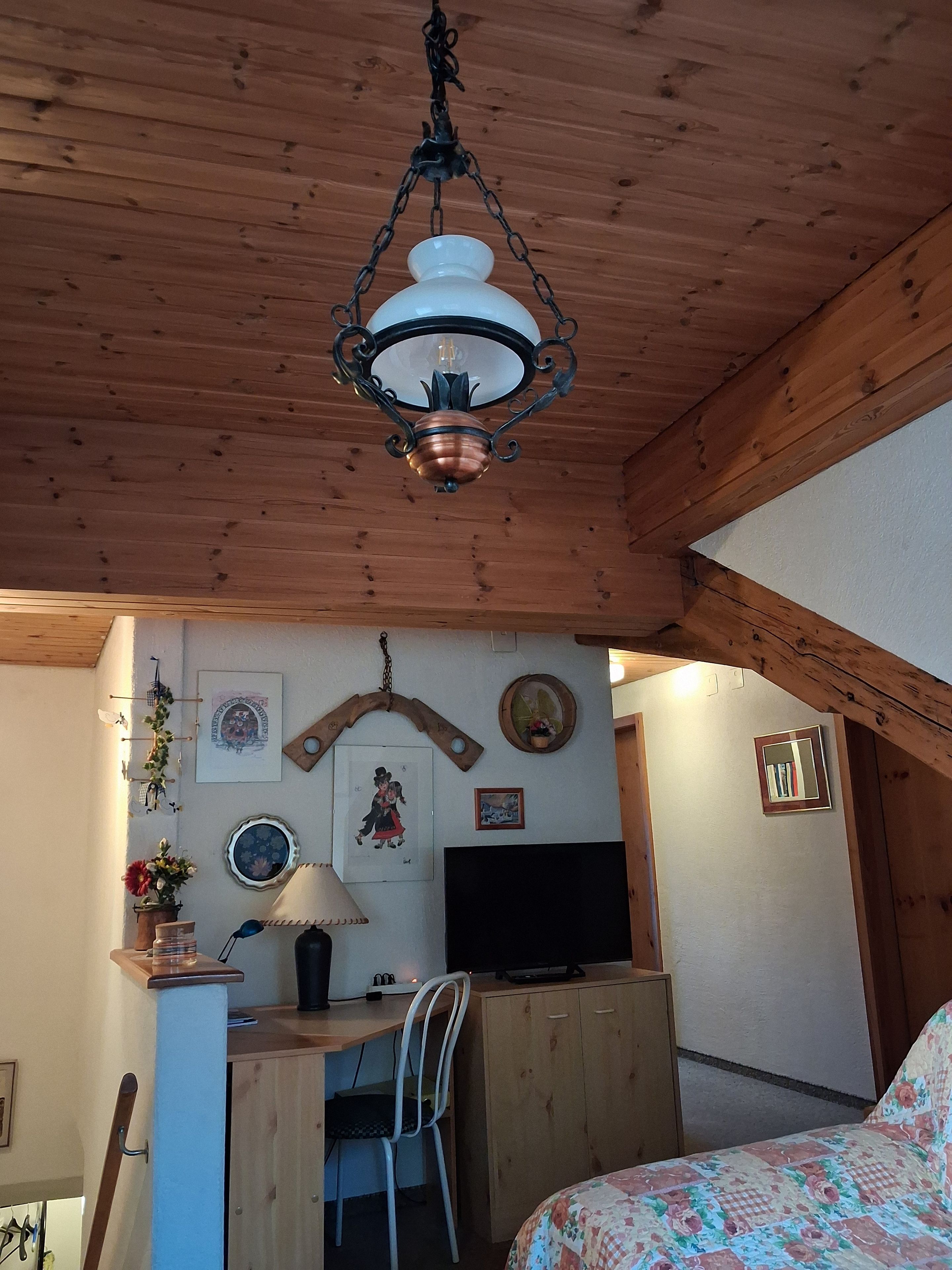 Una habitación acogedora con techo de madera, un escritorio con silla, una cama con colcha estampada y varias decoraciones en las paredes, incluyendo cuadros y un espejo.