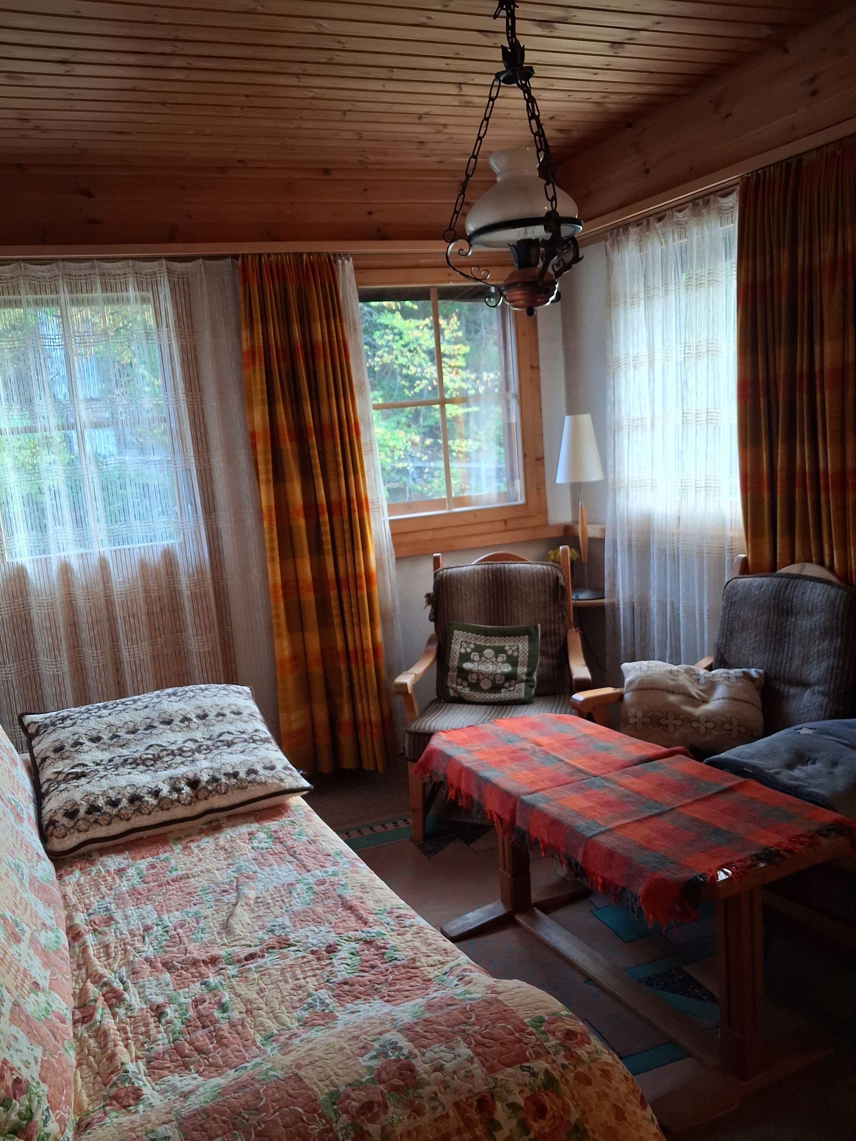 Una habitación acogedora con paredes de madera, una cama, un sofá y una mesa. Hay una ventana con cortinas, una lámpara colgante y una lámpara en una mesita de noche.