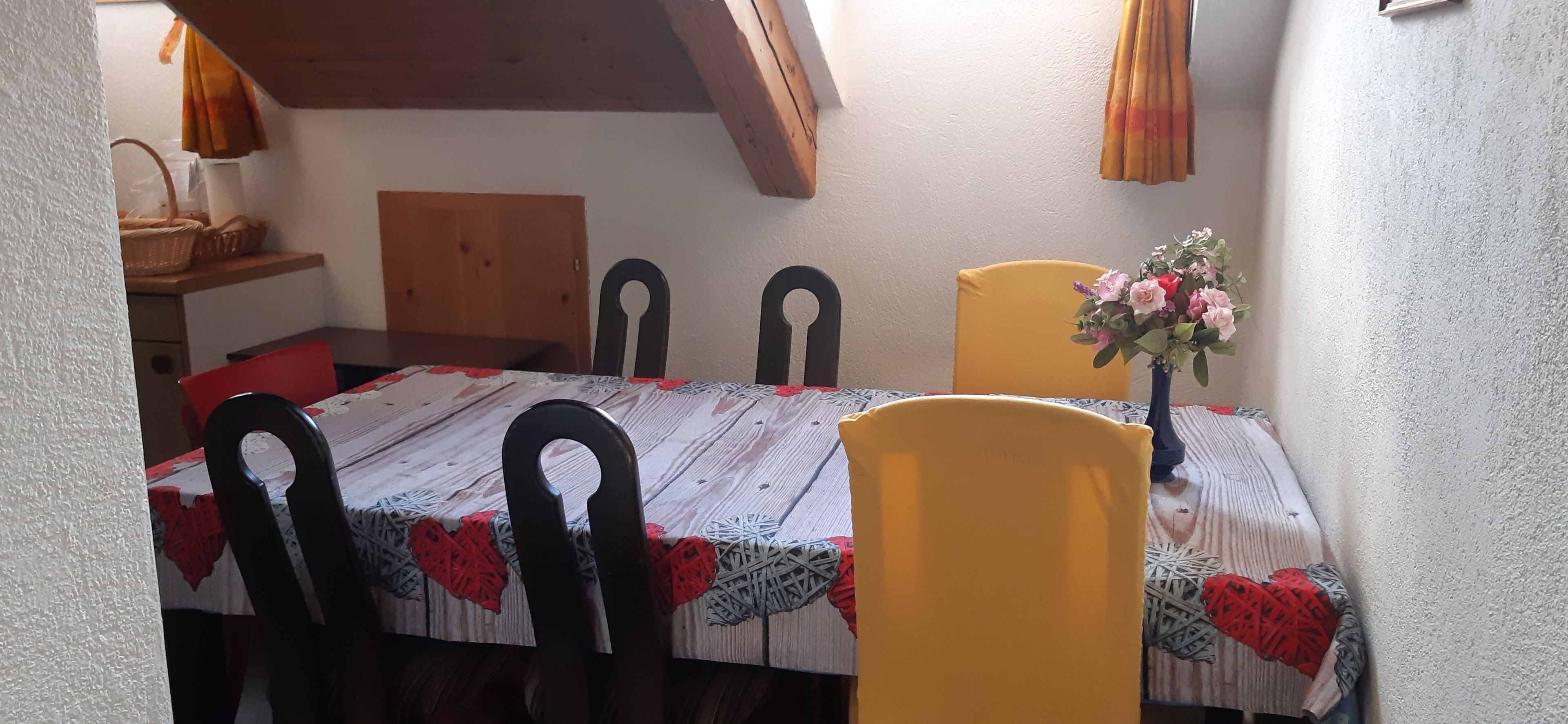 A dining area with a table set for four, wooden chairs, a vase with flowers, and a wooden headboard against the wall.