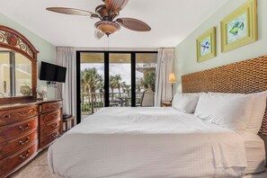 Een hotelkamer met een groot bed, een plafondventilator, een televisie, een dressoir en een balkon met uitzicht op palmbomen en een zwembad.