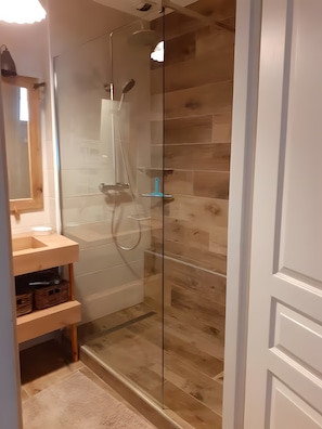 Eine gläserne Duschkabine, ein Holzboden, ein weißes Waschbecken mit Holzablage und ein Spiegel über dem Waschbecken.