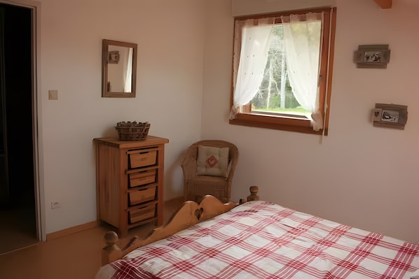 Ein Schlafzimmer mit einem Bett, einer Holzkommode, einem Sessel, einem Fenster mit Vorhängen und einem wandmontierten Spiegel.