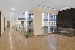 Amplio espacio interior con piso de madera, un sofá gris, plantas en macetas y una barandilla de vidrio con vista a un edificio de varios pisos.