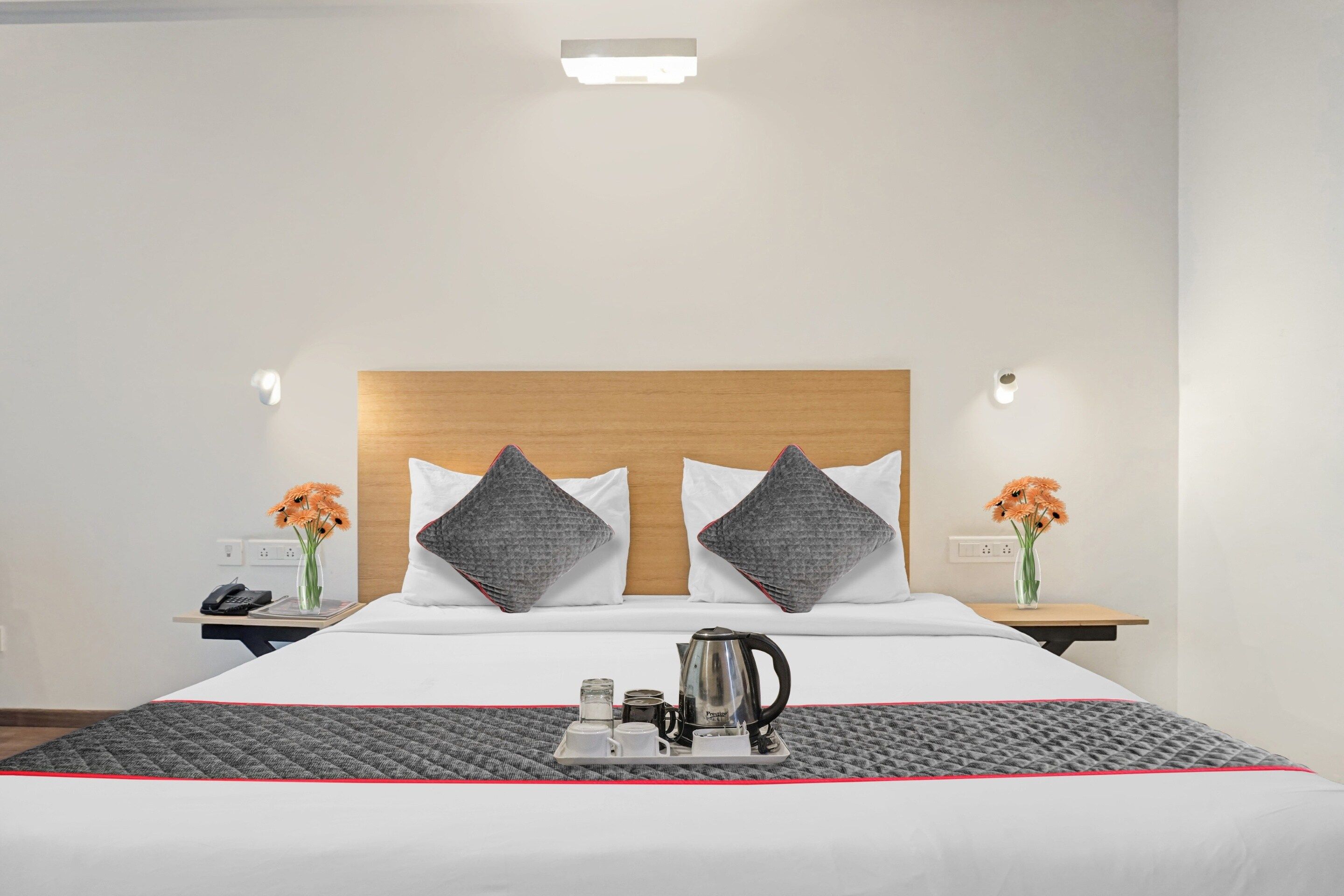 Una habitación de hotel con una cama, dos mesitas de noche, una mesita auxiliar, una tetera, una bandeja con tazas y un ceñidor, y dos jarrones con flores.