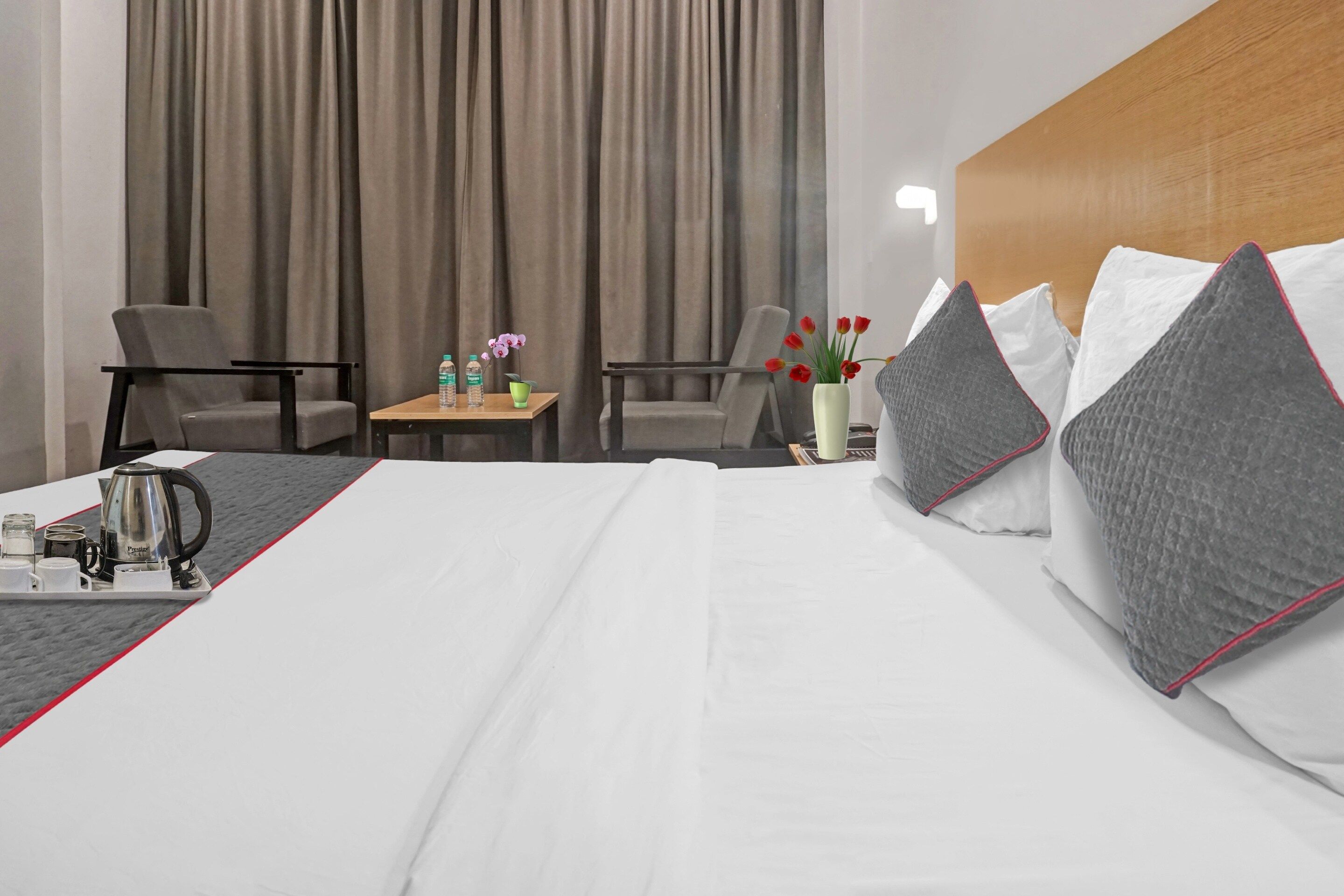 A hotel room with a bed, two chairs, a small table with a vase of flowers, and a tray with a kettle and cups.
