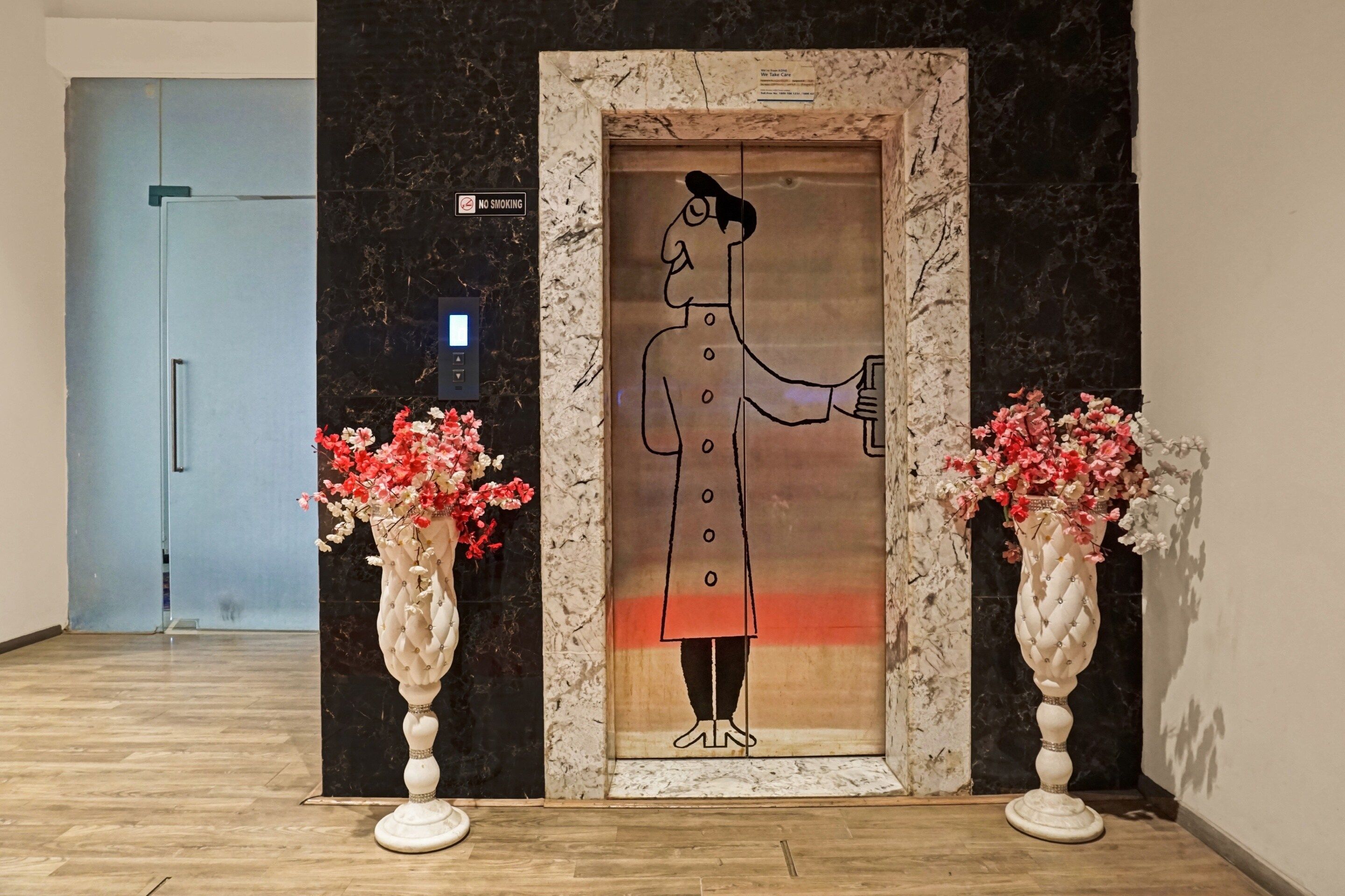 Un ascenseur dans lequel se trouve une personne tenant un téléphone, avec deux vases de fleurs de chaque côté, et un panneau « DÉFENSE DE FUMER » au-dessus.