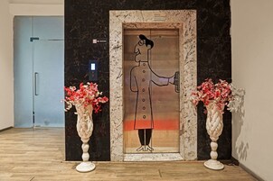 An elevator with a person holding a phone, flanked by two vases with flowers, and a 'NO SMOKING' sign above.