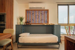 A compact bedroom with a bed, a small table, a chair, an air conditioning unit, and a window with a view. - Oceanfront 2-bedroom, 2.5-bath residence in El Zonte with floor-to-ceiling Pacific views, a sunset balcony, a n open kitchen, and access to Barefoot’s oceanfront pool, gym, yoga and massage rooms. Sleeps up to 6. (Chiltiupán)