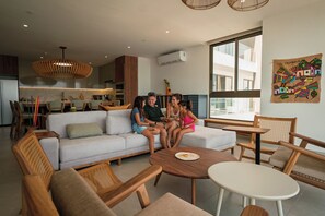 A modern living room with a sofa, chairs, a dining table, and a large window. - Oceanfront 2-bedroom, 2.5-bath residence in El Zonte with floor-to-ceiling Pacific views, a sunset balcony, a n open kitchen, and access to Barefoot’s oceanfront pool, gym, yoga and massage rooms. Sleeps up to 6. (Chiltiupán)