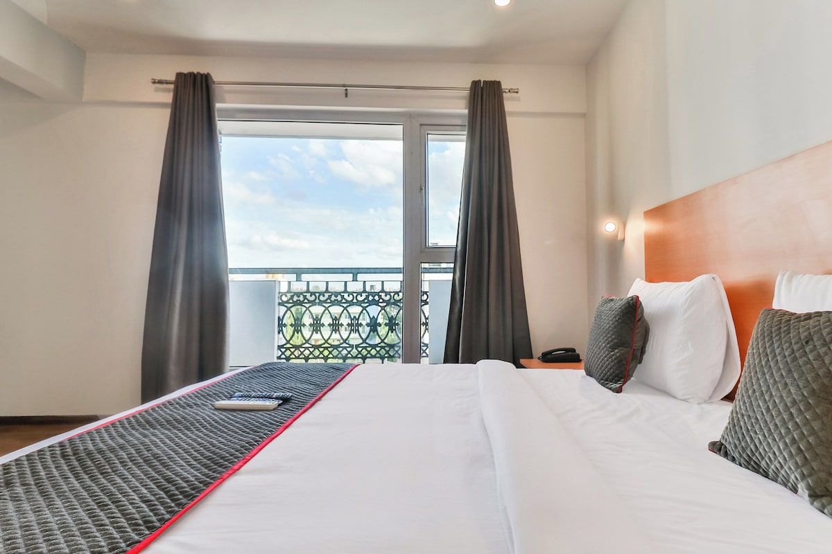 A hotel room with a bed, a window with curtains, a view of the outside, and a balcony with a railing.