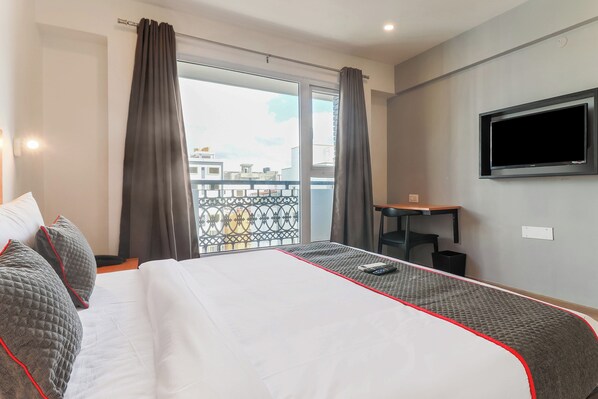 A hotel room with a bed, a TV mounted on the wall, a desk, and a balcony with a railing. - Townhouse Reva College University (Bangalore)