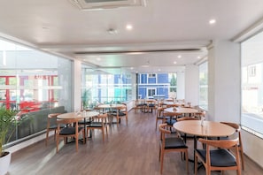 A modern dining area with multiple tables and chairs, large windows offering views of the outside, and a ceiling with recessed lighting. - Townhouse Reva College University (Bangalore)