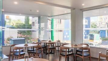 Un restaurante con mesas y sillas redondas, grandes ventanales que ofrecen vistas a edificios y zonas verdes.