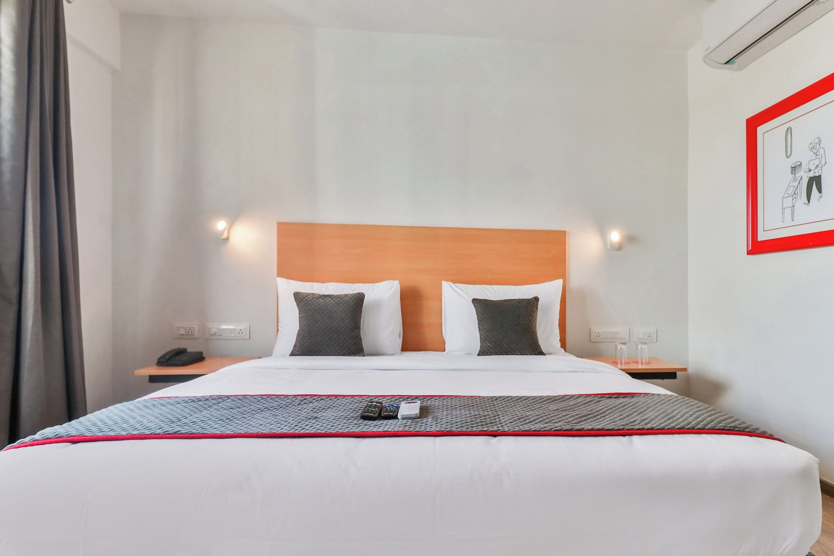 A hotel room with a bed, two bedside tables, a nightstand, a wall-mounted light, and a framed picture.