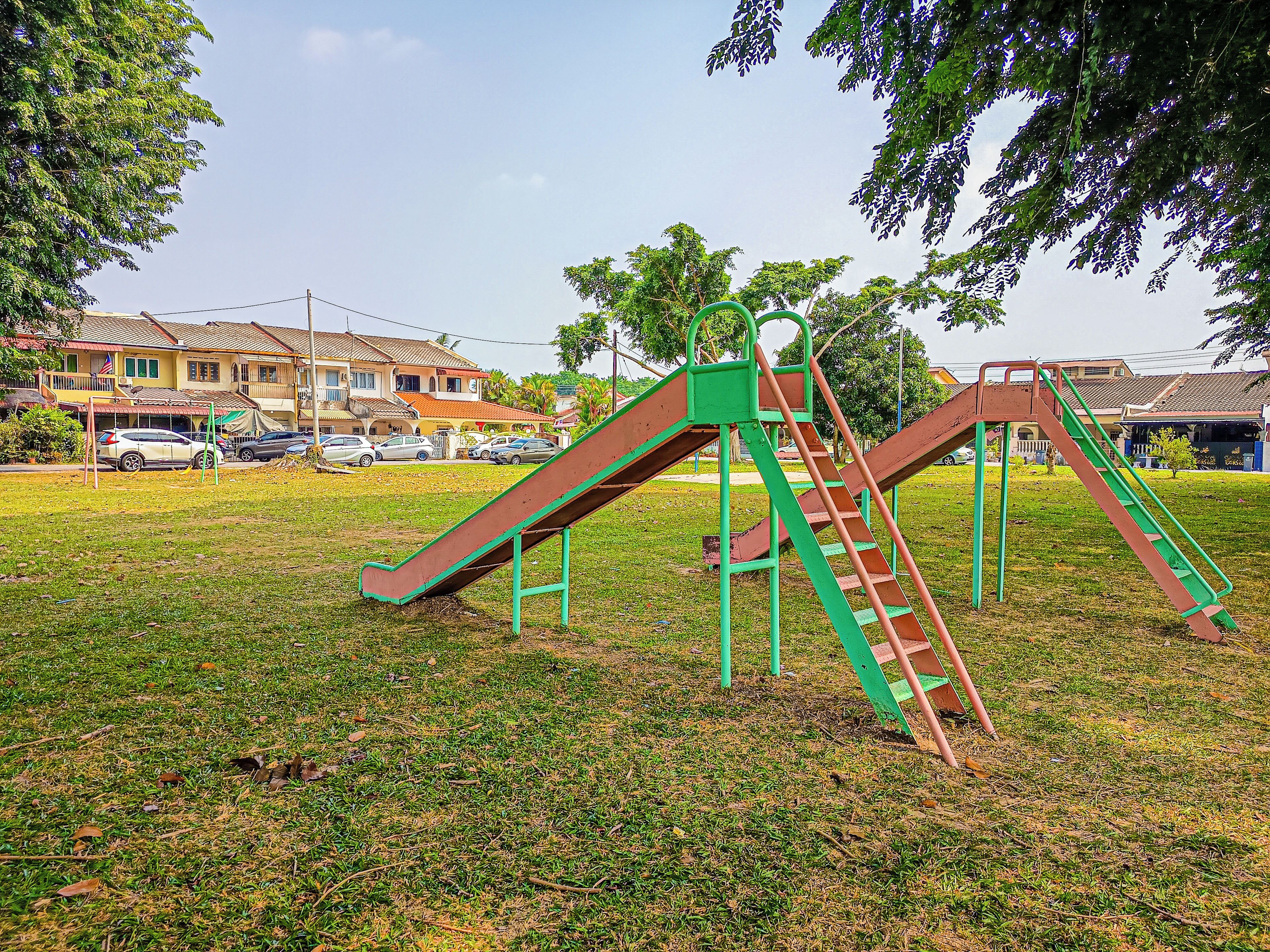 A playground with slides and climbing structures, surrounded by grass and trees, with houses in the background.
