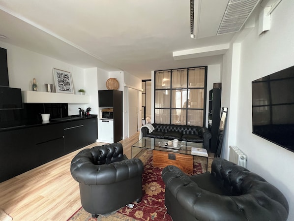Un espacio habitable moderno con un sofá de cuero negro, una mesa de centro de vidrio y una cocina con armarios negros y un microondas.