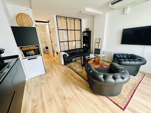 Una sala de estar moderna con un sofá de cuero negro, dos sillones de cuero, una mesa de centro de vidrio y un televisor de pantalla plana montado en la pared.