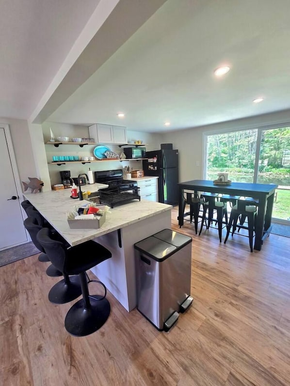 A modern kitchen with a central island, bar stools, a dining table with chairs, and a view of the outdoors.