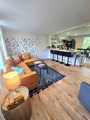 Ein modernes Wohnzimmer mit einer Couch, einem Couchtisch und einem gemusterten Teppich. Im Hintergrund ist eine Küchenzeile mit einer Bar und Hockern zu sehen.