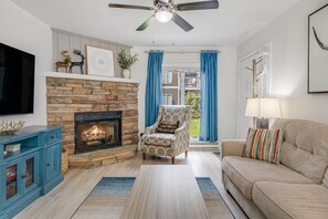 A living room with a stone fireplace, a ceiling fan, a television, a sofa, and a chair. - 1st Floor Condo | Walk to Main St | Fireplace (Blowing Rock)