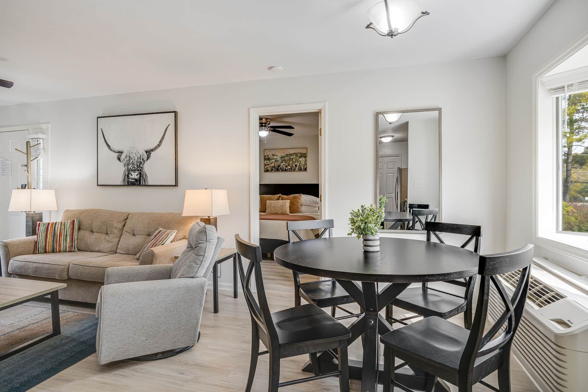 A modern living room with a sofa, armchair, and a dining table with chairs. There is a ceiling fan, a lamp, and a framed picture on the wall.