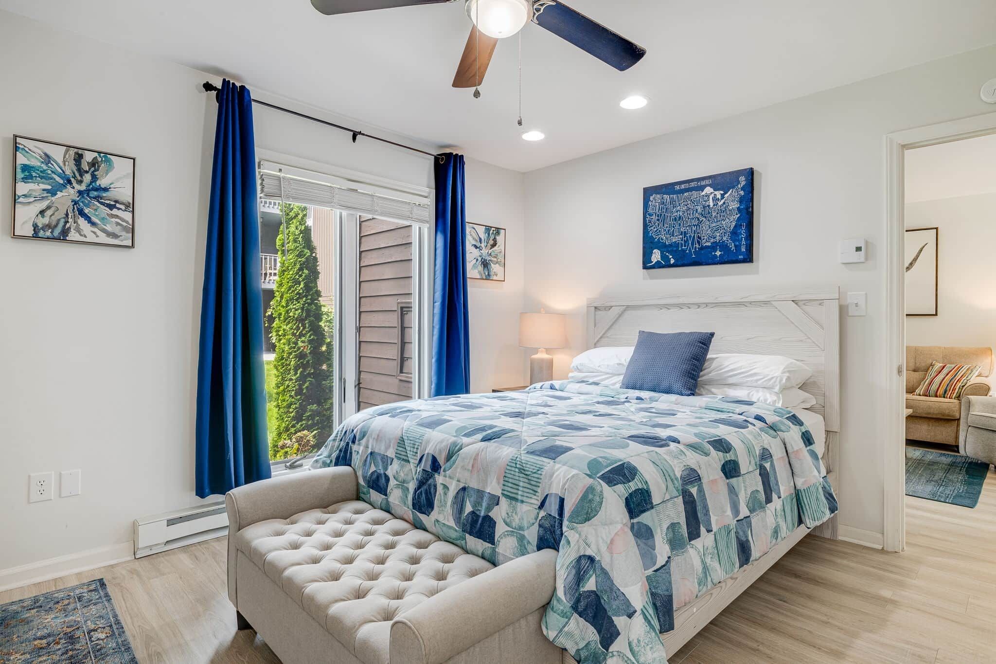 A bedroom with a bed, a ceiling fan, a window with curtains, and artwork on the walls.