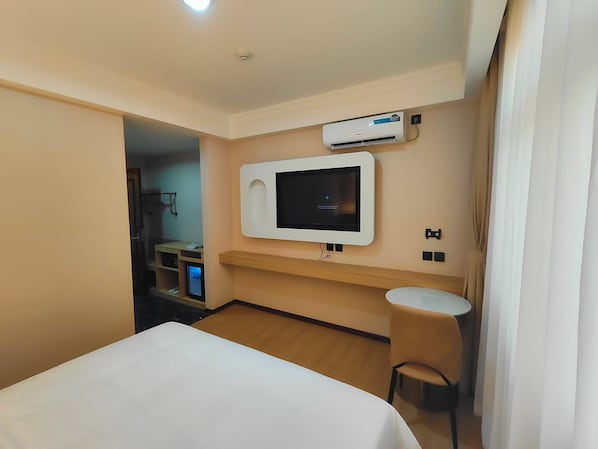 A hotel room with a bed, a TV mounted on the wall, a small table, and an air conditioning unit.