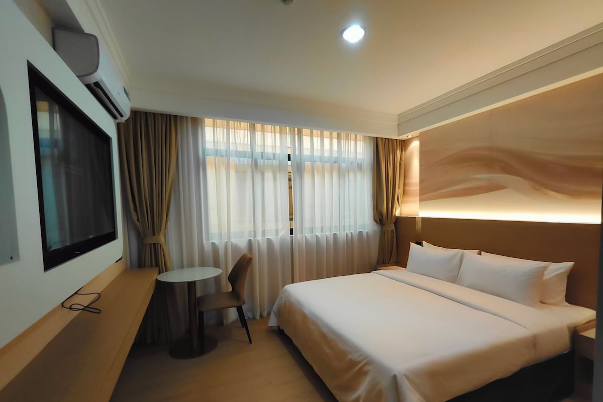 Quarto de hotel com uma cama, televisão na parede, mesa redonda pequena e uma cadeira.
