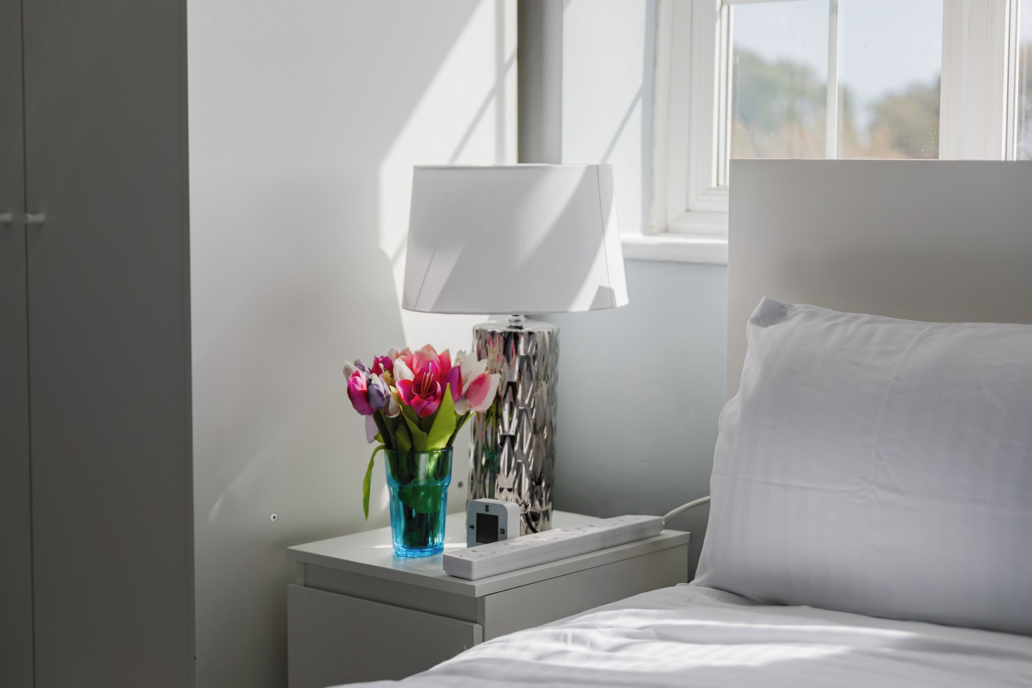 Una cama bien hecha con sábanas blancas, una mesita de noche con una lámpara y un florero con flores.