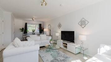 Una sala de estar moderna con un sofá blanco, una mesa de centro de vidrio, un televisor de pantalla plana y un área de comedor al fondo.