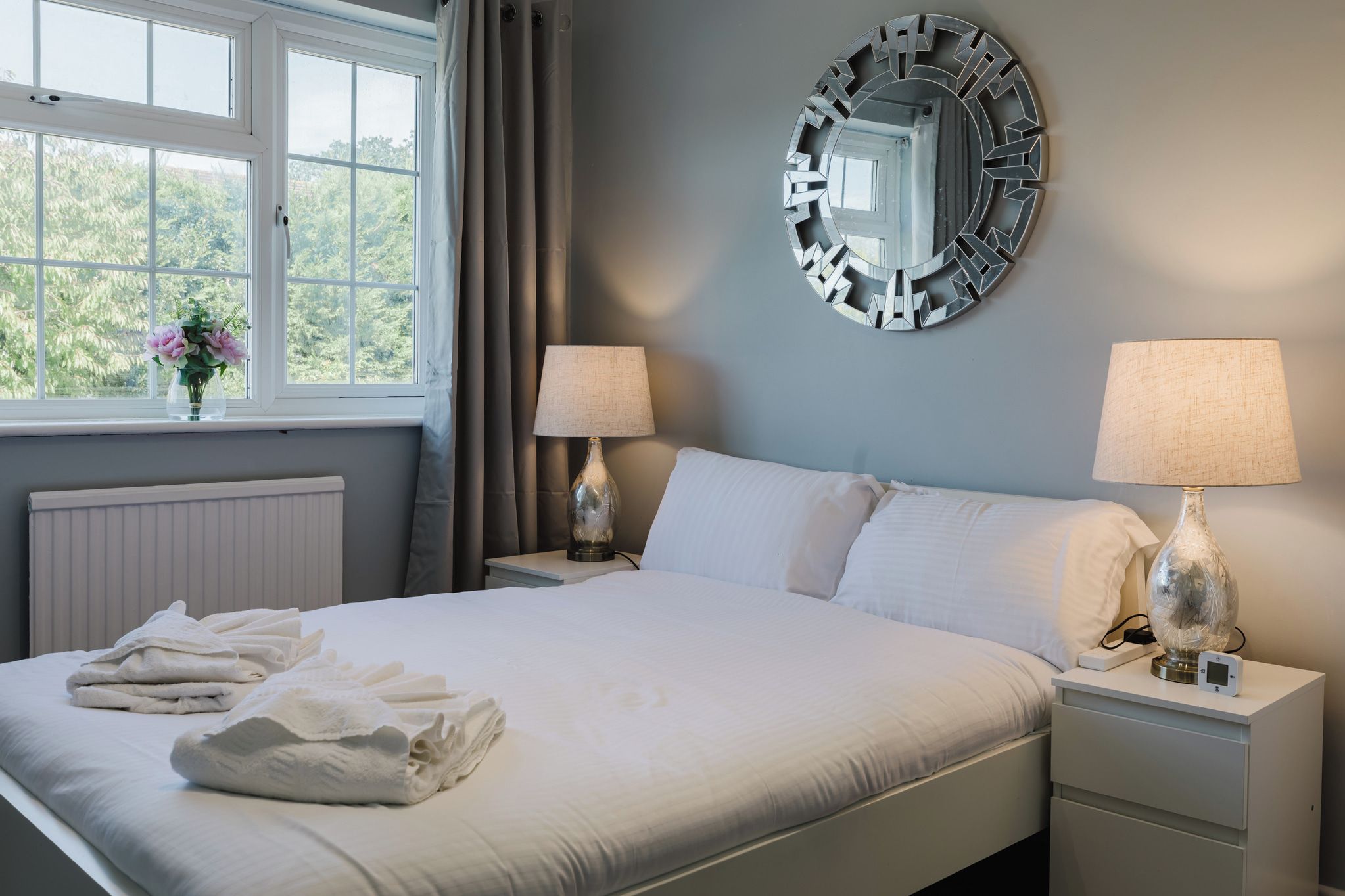 A neatly made bed with folded towels, two bedside lamps, a wall mirror, and a window with curtains.