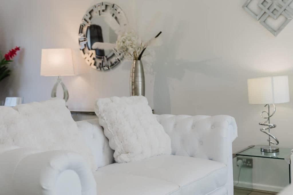 A white sofa with a fluffy pillow, a round mirror, a vase with flowers, and a glass table with a lamp.