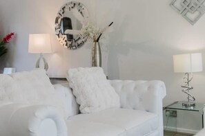 A white sofa with a fluffy pillow, a round mirror, a vase with flowers, and a glass table with a lamp.