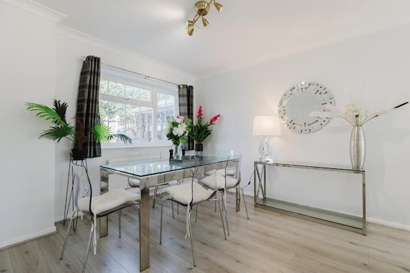 A dining area with a glass table and chairs, a potted plant, a vase with flowers, and a wall clock.