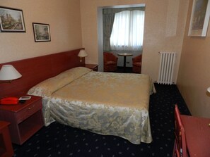 Habitació d'hotel amb un llit, una taula de treball, una cadira, un taulell i un radiador.