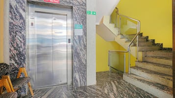 Un ascenseur doté d’un écran numérique, d’un panneau indiquant la sortie de secours en cas d’incendie et d’un escalier avec des rampes en métal.