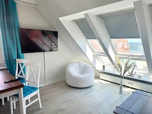 A modern living room with a white chair, a bean bag, a TV mounted on the wall, and a view of the city through large windows. - House With sea View - Apt. 4 (Wyk auf Foehr)