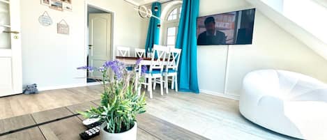 A modern living room with a white sofa, a dining area with blue chairs, a potted plant, and a TV mounted on the wall.