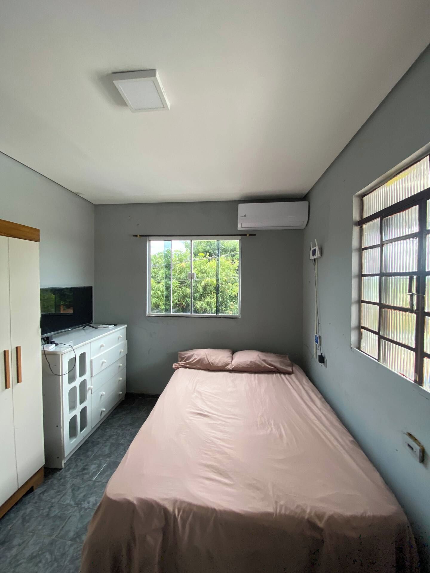 Ein Zimmer mit einem Bett, einem Fenster mit Blick auf Grünflächen, einem weißen Kleiderschrank und einer Klimaanlage.